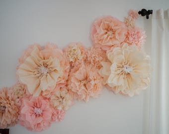 Könnte beinhalten: Eine Ansammlung großer Papierblumen in Pfirsich-, Rosa- und Cremetönen schmückt eine weiße Wand. Die Blumen variieren in Größe und Textur und bilden eine weiche, dekorative Darstellung. Rechts hängt ein weißer Vorhang.