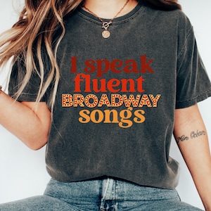 May include: A gray t-shirt with the text "I speak fluent BROADWAY songs" in red, orange, and white letters. The text is stylized to look like a marquee.