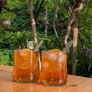 Könnte beinhalten: Zwei klare Trinkgläser gefüllt mit einer bernsteinfarbenen Flüssigkeit, Eiswürfeln, einer Orangenscheibe und einem Rosmarinzweig. Jedes Glas hat ein kreisförmiges, geprägtes Logo auf der Vorderseite und einen klaren Glasstrohhalm.