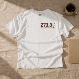 Puede incluir: Camiseta blanca con el texto "Z73.3 stress, not elsewhere classified" en marrón. Un pequeño gráfico de pato está junto al número. La camiseta está sobre una superficie neutra con una taza de café y un libro.