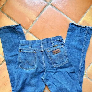 Puede incluir: Vaqueros Wrangler azules con un dise&ntilde;o cl&aacute;sico de cinco bolsillos. Los vaqueros presentan un parche de cuero marr&oacute;n con el logotipo de Wrangler en el bolsillo trasero. Los vaqueros est&aacute;n colocados sobre una superficie embaldosada.