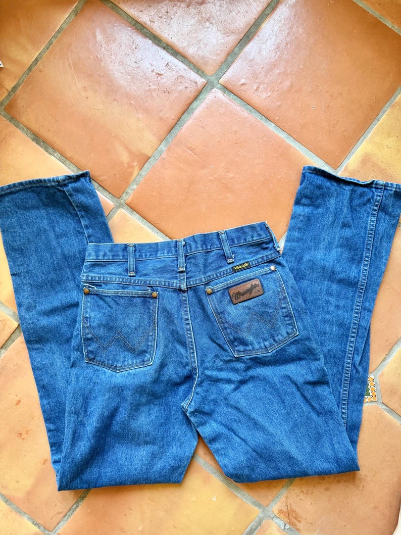 Puede incluir: Vaqueros Wrangler azules con un dise&ntilde;o cl&aacute;sico de cinco bolsillos. Los vaqueros presentan un parche de cuero marr&oacute;n con el logotipo de Wrangler en el bolsillo trasero. Los vaqueros est&aacute;n colocados sobre una superficie embaldosada.