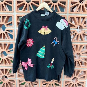May include: Black long-sleeved Christmas sweater with various festive appliques including a wreath, a bell, a poinsettia, a Christmas tree, candy canes, a candle, and Santa Claus.