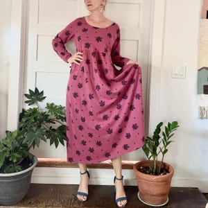 May include: A long-sleeved, mauve-colored dress with a scoop neckline and a flowing skirt. The dress is adorned with a pattern of dark purple maple leaves. The model is wearing blue sandals.