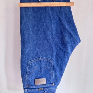 May include: A pair of medium-wash blue denim jeans hanging from a wooden hanger. The jeans feature a back pocket with a signature W-stitch pattern and a brown leather-like patch embossed with the Wrangler brand name and logo.
