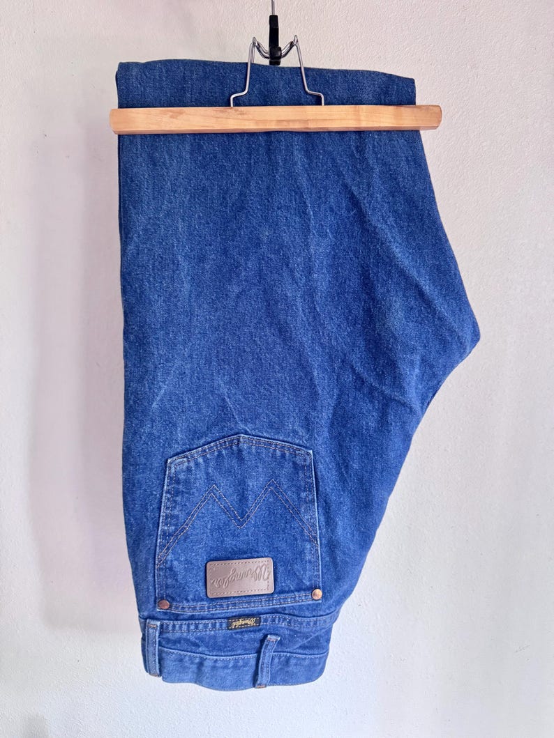 May include: A pair of medium-wash blue denim jeans hanging from a wooden hanger. The jeans feature a back pocket with a signature W-stitch pattern and a brown leather-like patch embossed with the Wrangler brand name and logo.