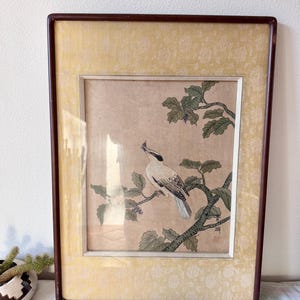 May include: Framed vintage artwork featuring a bird perched on a branch with green leaves and small purple berries. The bird is depicted in shades of white, gray, and black. The artwork is framed in a dark brown wooden frame with a yellow patterned border.