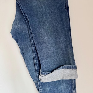 May include: Folded blue denim jeans with a cuffed hem. The jeans are a medium wash with a slightly faded appearance. The cuff reveals a lighter gray fabric.