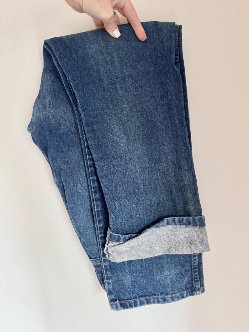 May include: Folded blue denim jeans with a cuffed hem. The jeans are a medium wash with a slightly faded appearance. The cuff reveals a lighter gray fabric.