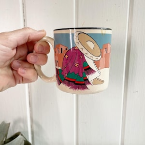 May include: A ceramic coffee mug with a colorful illustration of a person wearing a traditional Mexican outfit and a large hat. The background features a desert scene with buildings.