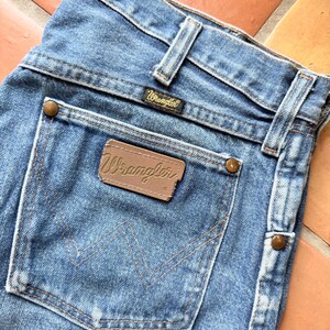 K&ouml;nnte beinhalten: Nahaufnahme einer hellblauen Jeans mit dem Wrangler-Logo auf der Ges&auml;&szlig;tasche und dem Bund. Die Jeans hat ein klassisches F&uuml;nf-Taschen-Design mit kupferfarbenen Nieten und einem braunen Lederaufn&auml;her.