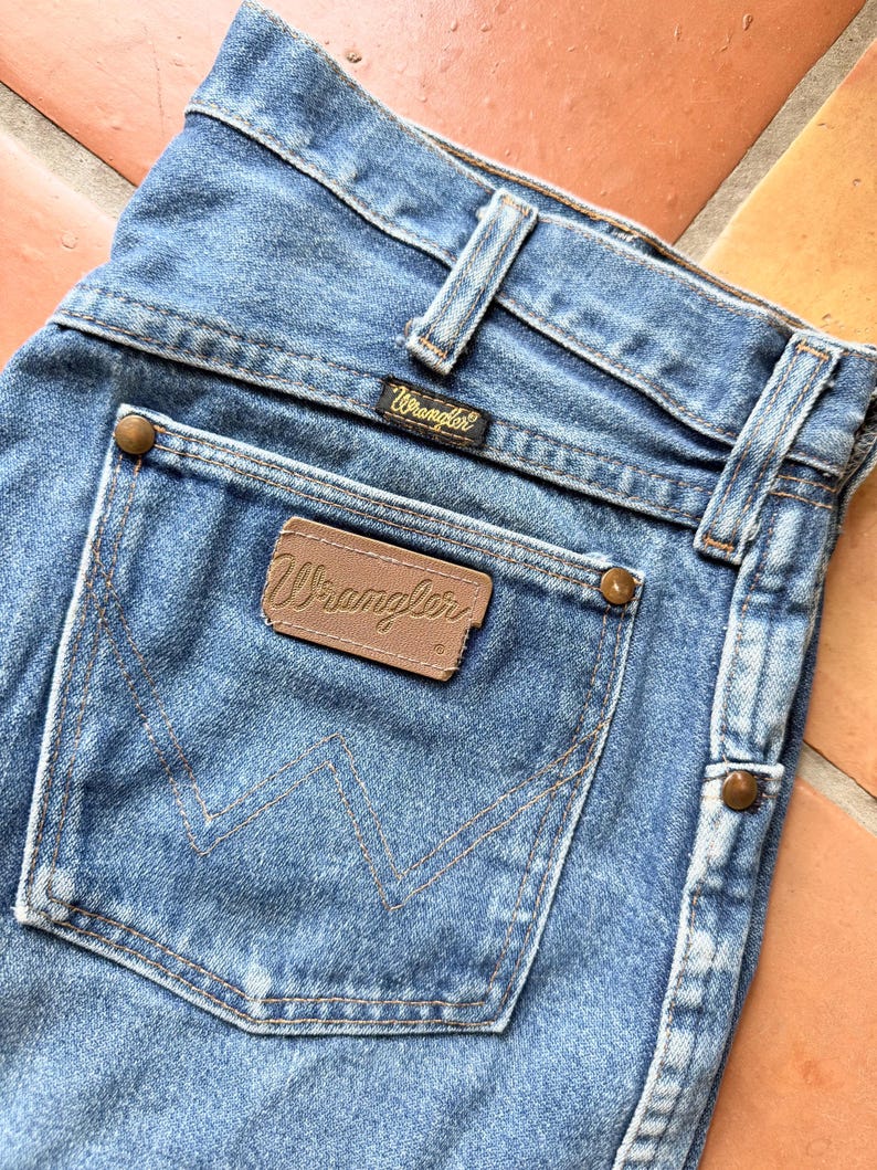 K&ouml;nnte beinhalten: Nahaufnahme einer hellblauen Jeans mit dem Wrangler-Logo auf der Ges&auml;&szlig;tasche und dem Bund. Die Jeans hat ein klassisches F&uuml;nf-Taschen-Design mit kupferfarbenen Nieten und einem braunen Lederaufn&auml;her.