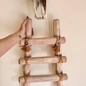 May include: A small, decorative wooden ladder painted in a distressed pink and white finish. The ladder has three rungs and is adorned with natural fiber accents and silver-toned metal embellishments. The ladder is approximately 18 inches tall.