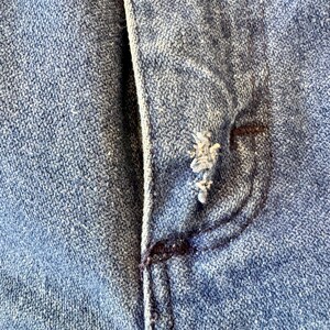 May include: Close-up of a section of blue denim fabric, showing the texture and stitching details. The image highlights a pocket and button area, with visible wear and tear, suggesting a vintage or distressed look.