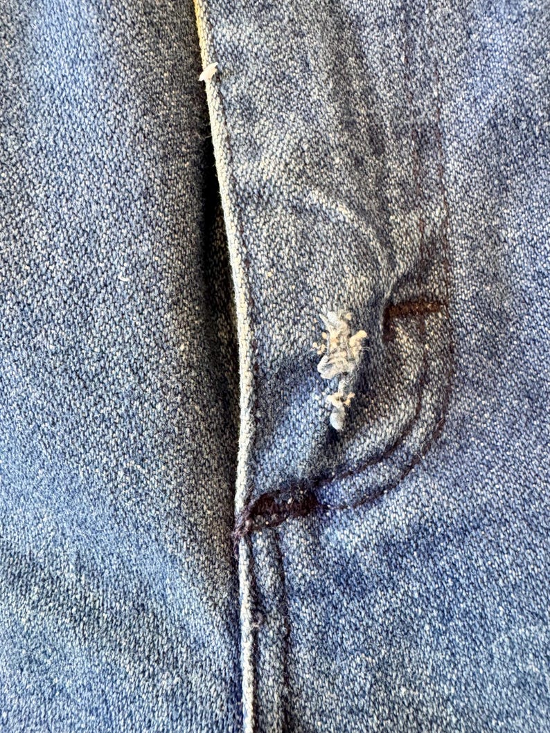 May include: Close-up of a section of blue denim fabric, showing the texture and stitching details. The image highlights a pocket and button area, with visible wear and tear, suggesting a vintage or distressed look.