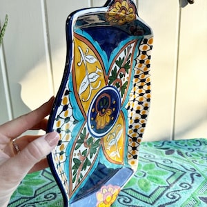 May include: A ceramic serving tray with a blue, yellow, green, and white floral design. The tray has a wavy edge and a handle on one side.
