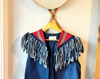 May include: A denim vest with fringe detailing around the neckline. The vest has a red and blue striped trim around the neckline.