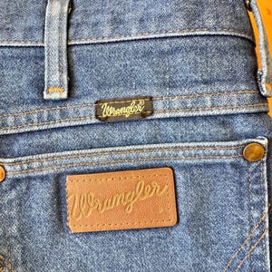 May include: Close-up of the back of blue denim Wrangler jeans. The jeans feature a brown leather patch with the Wrangler logo in a cursive font, and a black tag with the Wrangler logo. The stitching is in a contrasting orange color.