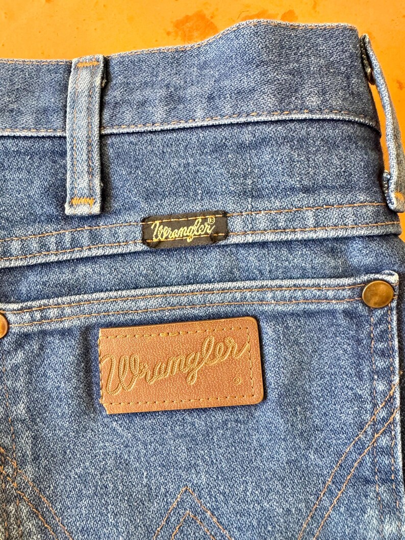 May include: Close-up of the back of blue denim Wrangler jeans. The jeans feature a brown leather patch with the Wrangler logo in a cursive font, and a black tag with the Wrangler logo. The stitching is in a contrasting orange color.
