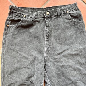 Op de afbeelding: Close-up van een zwarte denim jeans. De jeans heeft een klassiek ontwerp met vijf zakken en een knoop- en ritssluiting. De stof lijkt licht vervaagd, wat hem een vintage look geeft. De naden zijn wit.