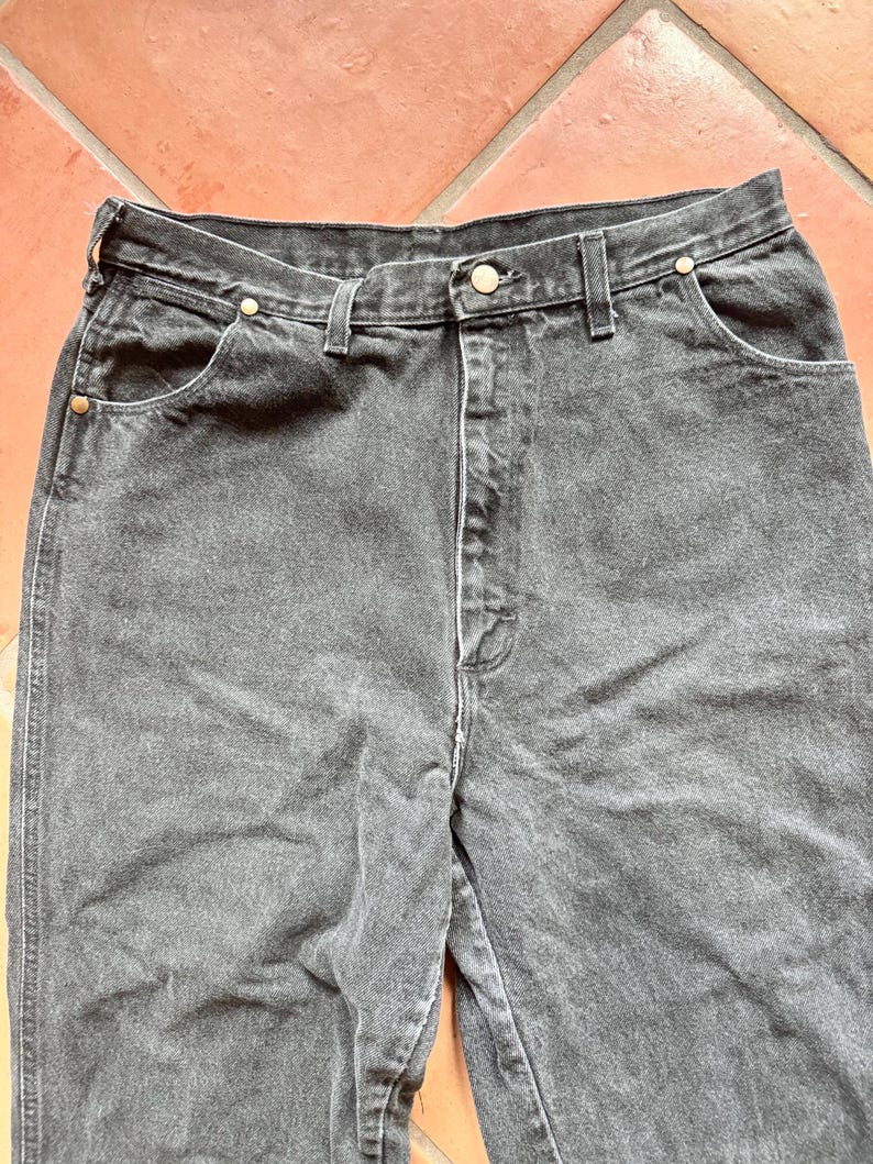 Op de afbeelding: Close-up van een zwarte denim jeans. De jeans heeft een klassiek ontwerp met vijf zakken en een knoop- en ritssluiting. De stof lijkt licht vervaagd, wat hem een vintage look geeft. De naden zijn wit.