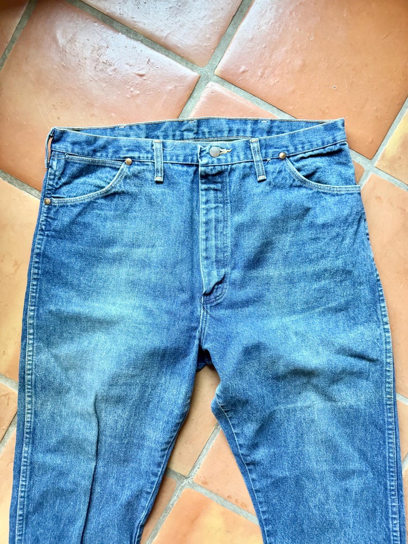 May include: Blue denim jeans with a classic five-pocket design. The jeans have a button and zipper closure, along with copper-colored rivets. The fabric shows signs of wear, suggesting a vintage or well-loved item.