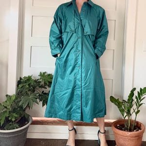 May include: Teal-colored trench coat with a button-up front, collared neckline, and long sleeves. The coat has pockets and a knee-length hem. The person is wearing blue heeled sandals. The coat is made of a shiny material.