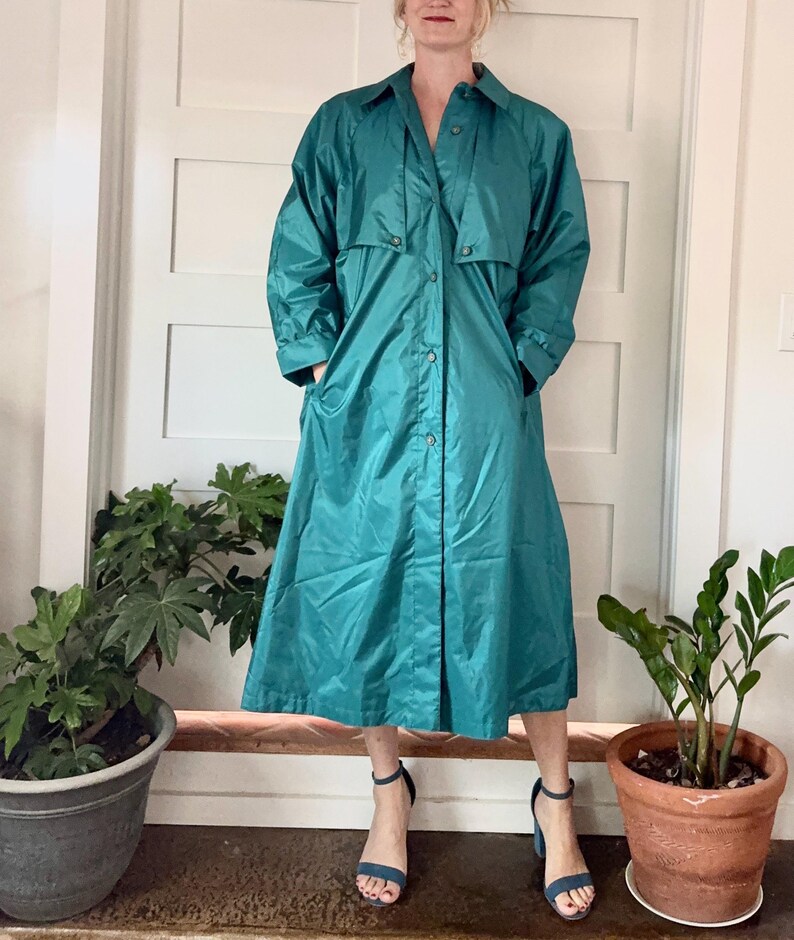 May include: Teal-colored trench coat with a button-up front, collared neckline, and long sleeves. The coat has pockets and a knee-length hem. The person is wearing blue heeled sandals. The coat is made of a shiny material.