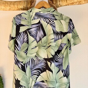 Peut inclure: Chemise &agrave; manches courtes avec un motif de feuilles tropicales. La chemise pr&eacute;sente de grandes feuilles vertes et vert clair avec des touches de violet sur un fond noir. Elle est suspendue &agrave; un cintre en bois.