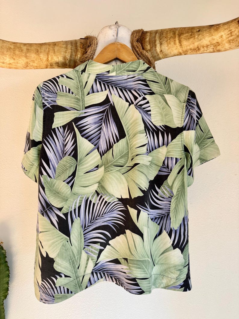 Peut inclure: Chemise &agrave; manches courtes avec un motif de feuilles tropicales. La chemise pr&eacute;sente de grandes feuilles vertes et vert clair avec des touches de violet sur un fond noir. Elle est suspendue &agrave; un cintre en bois.