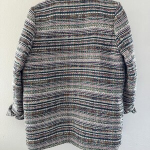 May include: A multi-colored tweed blazer with a collar and long sleeves. The jacket features a horizontal striped pattern in shades of brown, green, white, and black. The sleeves are cuffed at the wrists. The blazer is hanging on a wooden hanger.
