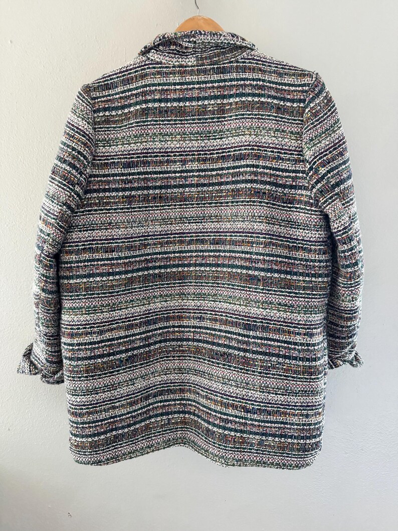 May include: A multi-colored tweed blazer with a collar and long sleeves. The jacket features a horizontal striped pattern in shades of brown, green, white, and black. The sleeves are cuffed at the wrists. The blazer is hanging on a wooden hanger.