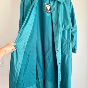 May include: Teal-colored, button-down raincoat with a collared neckline. The coat is open, revealing a darker teal lining and a tag with the brand name. The coat is hanging on a wooden hanger against a white wall.