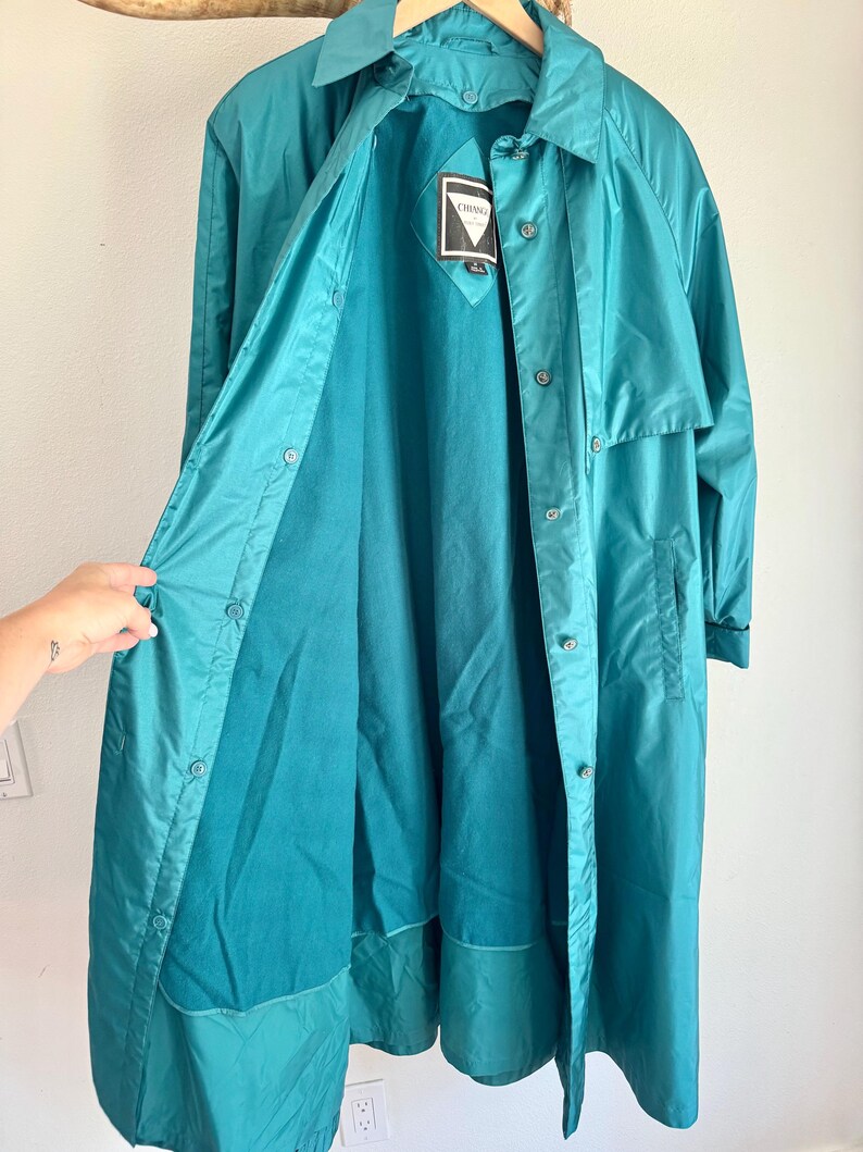 May include: Teal-colored, button-down raincoat with a collared neckline. The coat is open, revealing a darker teal lining and a tag with the brand name. The coat is hanging on a wooden hanger against a white wall.