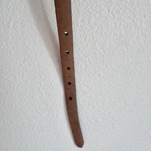May include: A close-up of a brown leather belt with several evenly spaced holes. The belt is a light brown colour and appears to be made of genuine leather. The background is a textured white wall.
