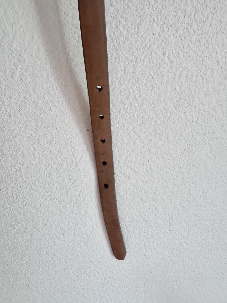 May include: A close-up of a brown leather belt with several evenly spaced holes. The belt is a light brown colour and appears to be made of genuine leather. The background is a textured white wall.