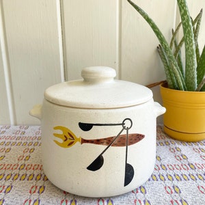May include: A white ceramic crock pot with a lid. The crock pot has a brown, black, and yellow design of a stylized bird with a fork in its beak.
