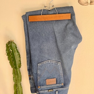May include: Blue denim jeans hanging on a wooden and metal hanger. The jeans have a classic five-pocket design, with a brown leather patch and the Wrangler logo on the back pocket. The jeans are folded neatly.