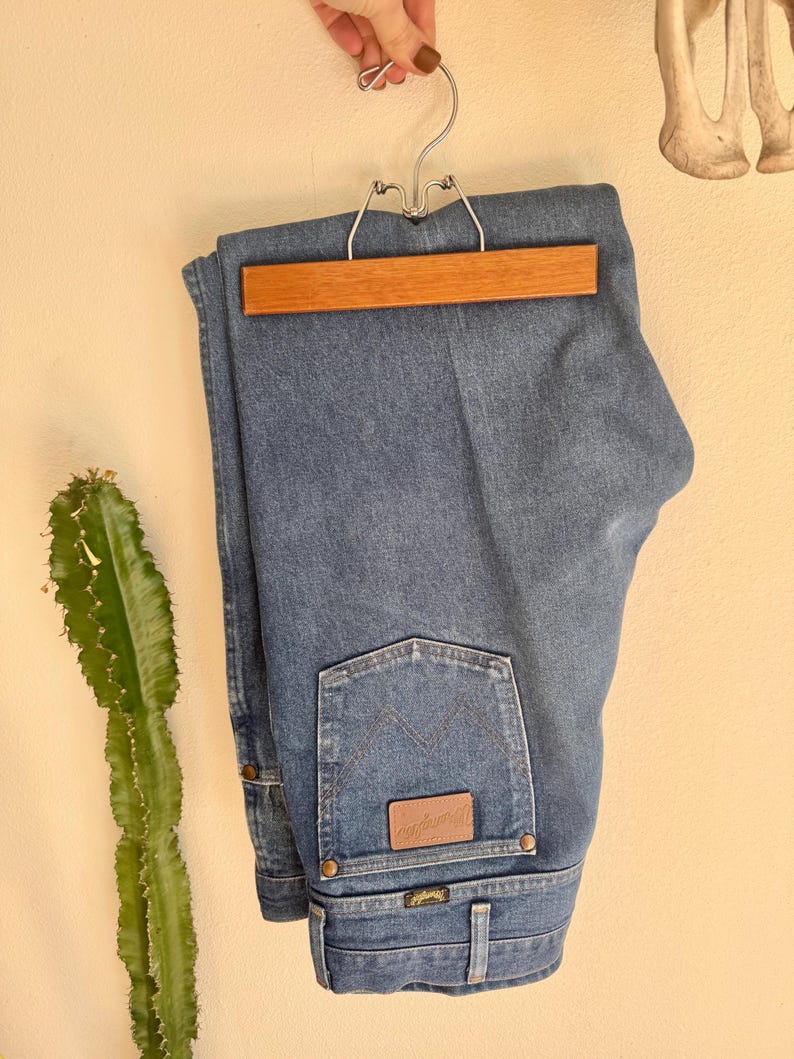 May include: Blue denim jeans hanging on a wooden and metal hanger. The jeans have a classic five-pocket design, with a brown leather patch and the Wrangler logo on the back pocket. The jeans are folded neatly.