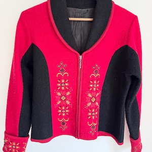 May include: A red and black wool jacket with a zip-up front and embroidered floral details. The jacket features a black collar and sleeves, with intricate embroidery in yellow, red, and gold. The cuffs also have matching embroidery.