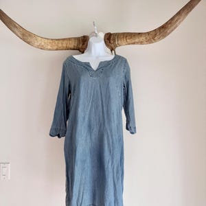 May include: A light blue denim dress with a v-neckline and three-quarter sleeves. The dress is displayed on a white mannequin against a white wall, with large, curved horns in the background.