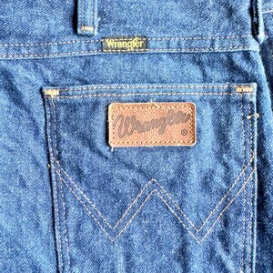K&ouml;nnte beinhalten: Nahaufnahme der Ges&auml;&szlig;tasche einer blauen Jeans. Die Tasche hat einen braunen Lederaufn&auml;her mit dem Markennamen "Wrangler" in goldfarbenen Buchstaben. Die Jeans hat ein klassisches Design mit sichtbaren N&auml;hten.