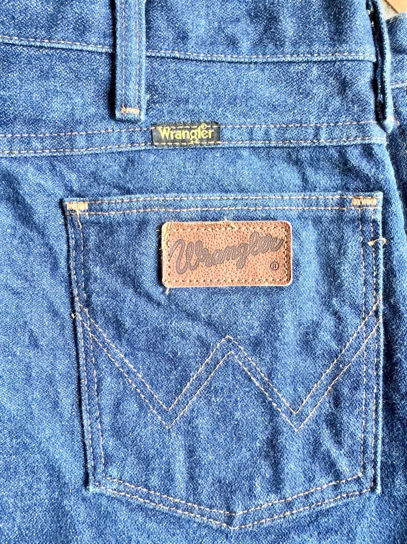 K&ouml;nnte beinhalten: Nahaufnahme der Ges&auml;&szlig;tasche einer blauen Jeans. Die Tasche hat einen braunen Lederaufn&auml;her mit dem Markennamen "Wrangler" in goldfarbenen Buchstaben. Die Jeans hat ein klassisches Design mit sichtbaren N&auml;hten.