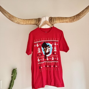 May include: Red short-sleeve t-shirt with a festive design. The graphic features a cartoon beaver wearing a hat and scarf, surrounded by snowflakes and trees. The shirt is hanging on a wooden hanger.