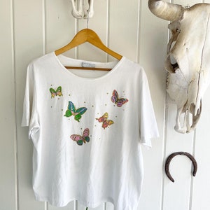 May include: White t-shirt with a colorful butterfly design. The butterflies are embroidered with sequins and beads.