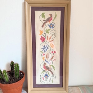 May include: A framed cross-stitch artwork featuring two birds and floral designs in shades of red, blue, orange, and green. The artwork is framed in a light brown wooden frame with a purple matting. A small cactus in a terracotta pot is in the foreground.