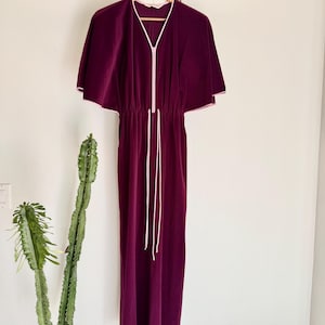 May include: A plum-colored velvet dress with a cape-like sleeve design. The dress features a white zipper and drawstring, with white trim along the neckline and sleeves. The dress is hanging on a wooden hanger.