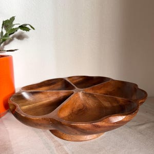 May include: A wooden serving dish with six sections, ideal for snacks or appetizers. The dish has a flower-like shape with a raised base. The wood has a warm, brown tone, and the dish is empty.