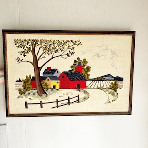 May include: A framed needlepoint embroidery depicting a rural scene with a red barn, yellow house, and a green tree. The scene is set against a backdrop of a blue sky with white clouds and a distant mountain range.
