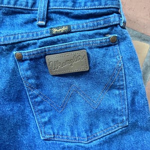Puede incluir: Vaqueros de mezclilla azul medio con un parche rectangular de cuero marr&oacute;n en el bolsillo trasero que muestra el logotipo de Wrangler en cursiva. El bolsillo incluye remaches met&aacute;licos de color cobre y una costura decorativa en forma de W.